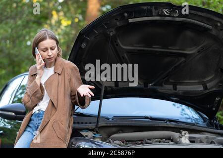 Helpless woman driver calling for help/assistance looking at broken down car, stopped at the roadside. Doesn't understand what happened. Stock Photo