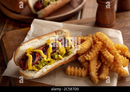 Grilled hot dog with mustard and relish on a bun and french fries on a rustic wooden board Stock Photo