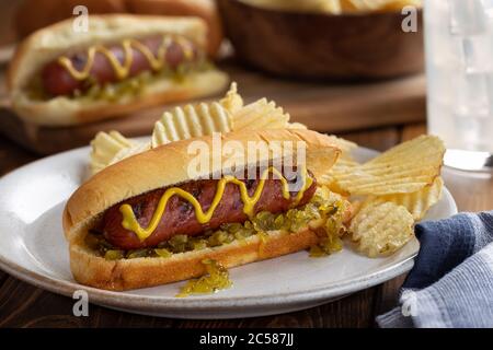 Grilled hot dog with mustard and relish on a bun and potato chips on a white plate Stock Photo