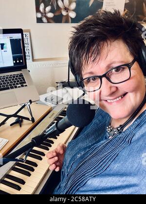 Mature female with black short hair & glasses smiling behind microphone, musical keyboard and computer whilst teaching online through livestreaming Stock Photo