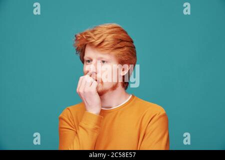 Nervous young redhead man in orange sweater biting fingernails against blue background Stock Photo