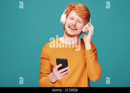 Portrait of positive handsome young redhead man with beard adjusting wireless headphones while choosing music track on smartphone Stock Photo
