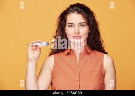 Portrait of young curly-haired woman showing her temperature on electronic thermometer against orange background, health and coronavirus concept Stock Photo