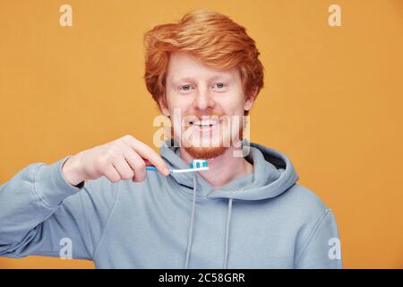 Portrait of positive young redhead man in hoodie cleaning teeth with toothbrush against orange background Stock Photo