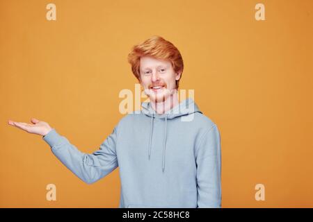 Portrait of content young red-bearded blogger in hoodie pointing aside while doing ads against orange background Stock Photo
