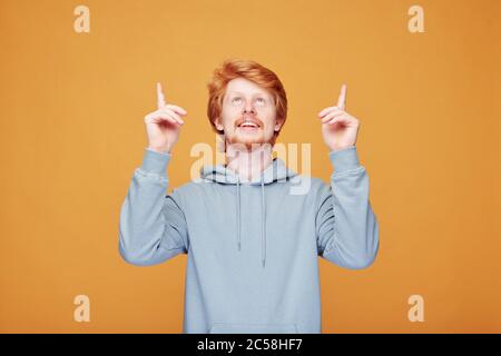 Positive young redhead man with beard standing against orange background and pointing up while telling about promotion Stock Photo