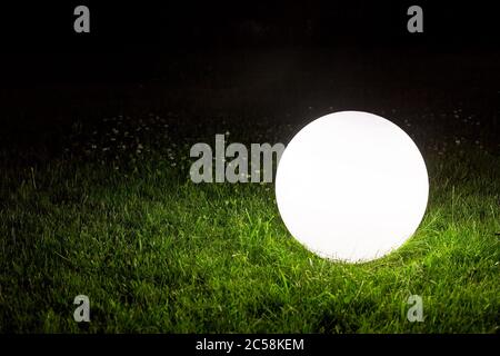 illuminating a spherical ground garden lamp of white color lies on a green lawn in the grass in the backyard of the park, closeup of a lighting fixtur Stock Photo