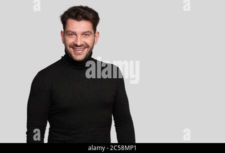 Smiling bearded young male model dressed casually, isolated over white background. Positive pleased guy being in good mood. Man in black turtleneck Stock Photo
