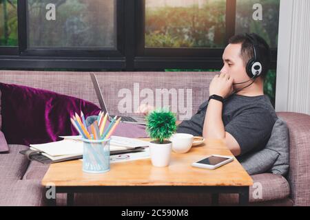 Young man lying on the couch and video call with colleagues by laptop in home office. Man wear headphone video calling or distance education e-leaning Stock Photo