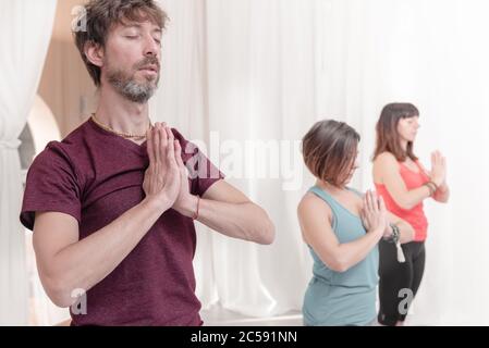 The man in the foreground with the hands in Namaste's gesture. Close up of man meditating at yoga studio. How to Keep Your Arms in the Air for a Kriya Stock Photo