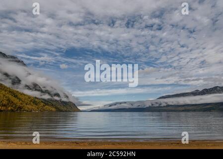 Amazing autumn view with a lake and mountains in fluffy clouds and fog against a blue sky and beautiful clouds Stock Photo