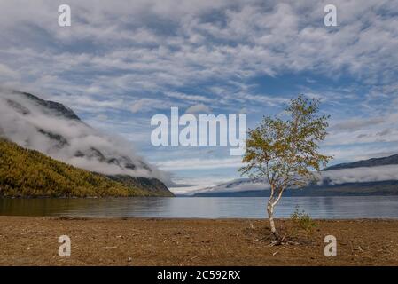 Amazing autumn view with a lake and mountains in fluffy clouds and fog against a blue sky and beautiful clouds Stock Photo