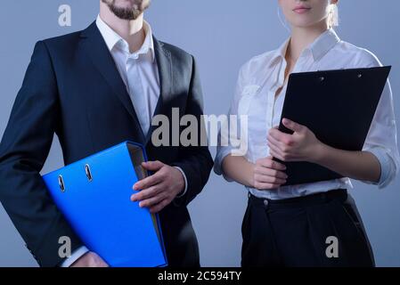 Portrait of young people, businessmen smiling, stand in business suits, holding office folders for papers in their hands. Studio photo on a gray Stock Photo