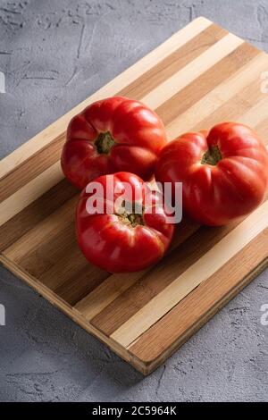 Three pink heirloom tomato vegetables, fresh red ripe tomatoes on wooden cutting board, vegan food, stone concrete background, angle view Stock Photo
