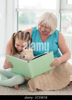 Cute granny reading book to little granddaughter holding toy sitting on floor in playroom Stock Photo
