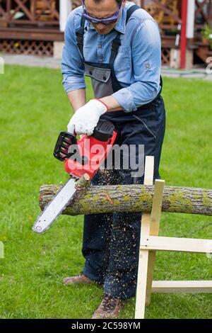 man in overalls saws wood with chain saw using sawhorse on green lawn Stock Photo