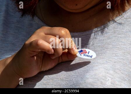 Shiloh, IL--June 26, 2020; female voter places red white and blue oval I Voted sticker on shirt with right hand after casting ballot Stock Photo