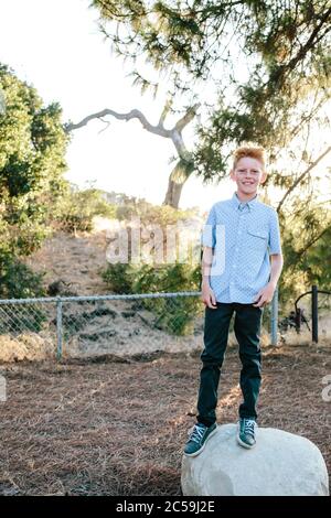 A Red Haired Boy Smiles While Standing On A Large Rock Stock Photo