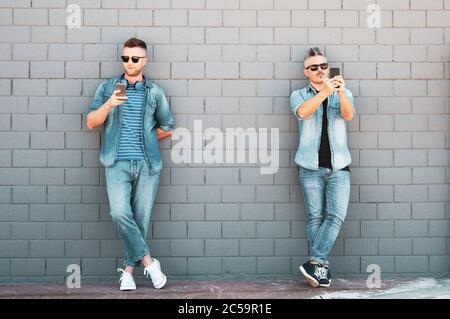 Two young man looking at mobile phone lean against brick wall - Couple of cool guys using smartphone standing outdoor in the street Stock Photo