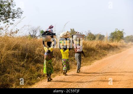 Women are walking with a bucket on their head walk on a dirt road in northern Benin. A woman carries her baby on her back. Stock Photo