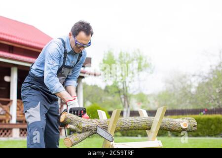 man in overalls saws wood with chain saw using sawhorse Stock Photo