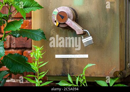 A rusty control box with a hand wheel and a padlock labelled "Kran Blech" (crane sheet metal) hangs on a brick wall of the Henschel-Hallen in Kassel, Germany – the disused works site of former locomotive and armaments manufacturer Henschel & Sohn. A sign below refers to the key held at the guardhouse; nettles grow in front of it. Stock Photo