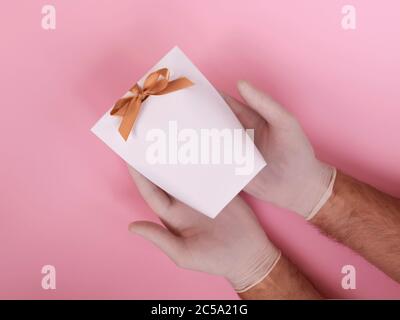 Male hands in medical glove holding a white gift bag on a pink background. Contactless giving a present during the quarantine period. Stock Photo