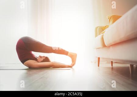 Young Woman enjoying evening yoga exercises doing Halasana pose at home living room near the big window. Active people and healthy lifestyle concept. Stock Photo