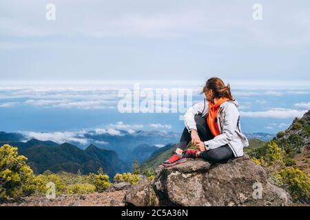 Young adult caucasian female dressed  sporty clothes resting on the rock and enjoying clouds view from Pico Ruivo mount 1861m - the highest peak on th Stock Photo