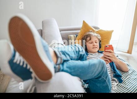 Preteen boy lying at home on cozy sofa dressed casual jeans and new sneakers listening to music and chatting using wireless headphones connected with Stock Photo