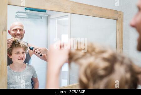 Bald dad and his long-haired teenager son in bathroom in front of the mirror. Father trying to trim long hairs showing to boy his new style haircut. C Stock Photo