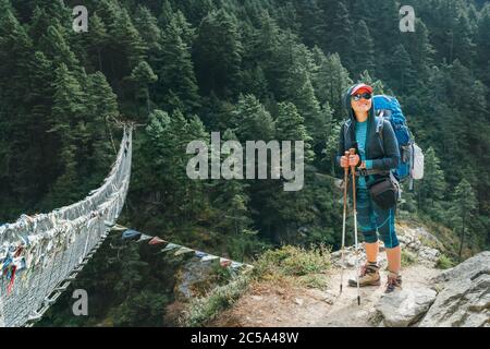 Young smiling female photographer walking Everest Base Camp route at canyon over Suspension Hillary Bridge with multicolored Tibetan Prayer flags hing Stock Photo
