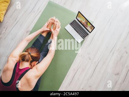 Fit sporty healthy woman sitting on mat in Paschimottanasana pose, doing breathing exercises, watching online yoga class on laptop computer. Healthy p Stock Photo