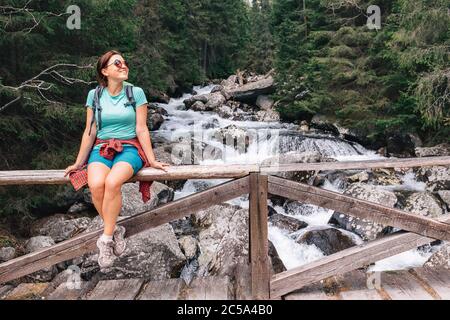 Young female backpacker resting on the mountain cold water stream bridge. Woman sitting on bridge fence smiling and enjoying the relaxing moment. Acti