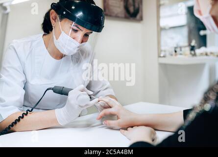 Professional manicure master in Transparent Safety Face Shield using Electric Nail Polisher Tool for Glazing treatment manicure procedure. Small busin Stock Photo