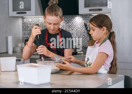Brother and sister dressed aprons making a homemade pancakes on the home kitchen. Boy using electric mixer for liquid dough mixing in the bowl and smi Stock Photo