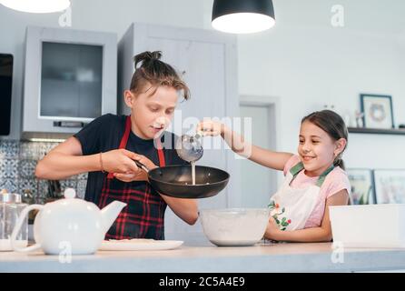 Sister and Brother dressed aprons making a homemade pancakes on the home kitchen. Girl poring a liquid dough on the hot pan. Kids Home cooking concept Stock Photo