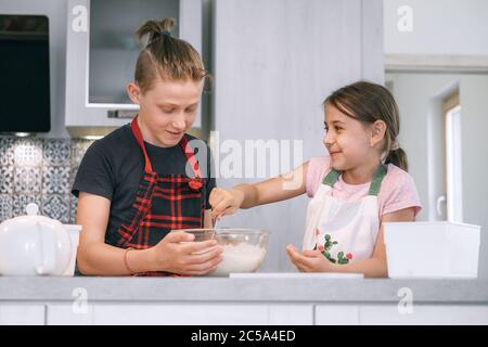 Brother and sister dressed aprons making a homemade pancakes on the home kitchen. Girl mixing a liquid dough in the bowl and smiling to brother. Kids Stock Photo
