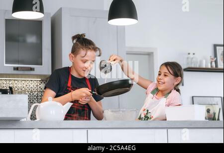 Kids Home cooking concept image. Sister and Brother dressed aprons making a homemade pancakes on the home kitchen. Girl poring a liquid dough on the h Stock Photo