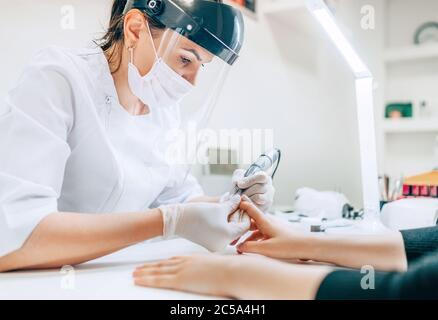 Small business existence at COVID-19 lockdown concept. Professional manicure master in Transparent Safety Face Shield using Electric Nail Polisher Too Stock Photo