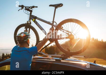 Young Man dresses modern cycling clothes and protective helmet installing his mountain bike on the car roof with sunset backlight. Active sporty peopl Stock Photo