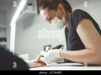 Professional manicure master dressed black in safety face mask using Electric Nail Polisher Tool for Glazing treatment manicure procedure. Everyday wo Stock Photo