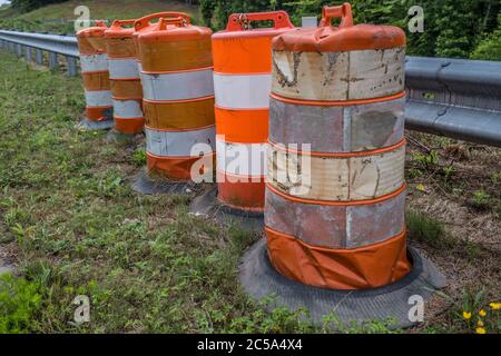 Several orange construction barrels sitting alongside a guardrail all tattered and worn waiting to be used again by the highway closeup Stock Photo