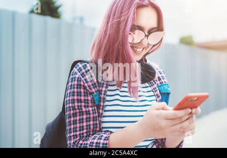 Portrait of Beautiful modern young smiling female teenager with extraordinary hairstyle color in a checkered shirt browsing an internet via smartphone Stock Photo