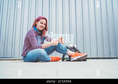 Beautiful modern smiling young female teenager in a checkered shirt and jeans with headphones and smartphone sitting on the street sidewalk. Modern te Stock Photo