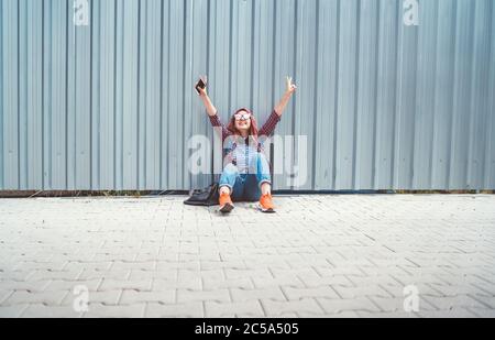 Cheerful hands rising UP Beautiful modern smiling young female teenager in a checkered shirt and jeans with headphones and smartphone sitting near a w Stock Photo