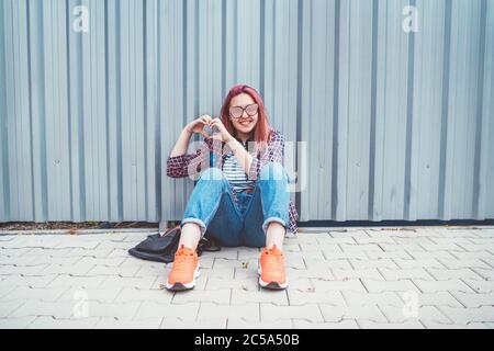Cheerful Beautiful modern smiling young female teenager in a checkered shirt and jeans with headphones sitting near a wall on the street and making a Stock Photo