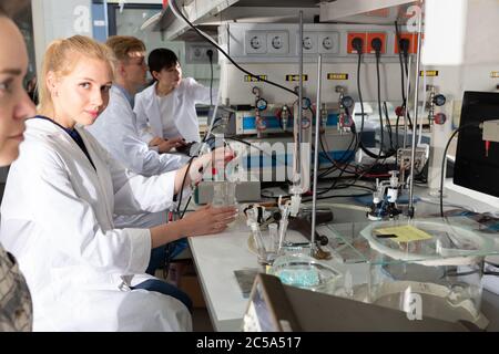 Portrait of young female lab technician engaged in research in chemical laboratory with her colleagues Stock Photo