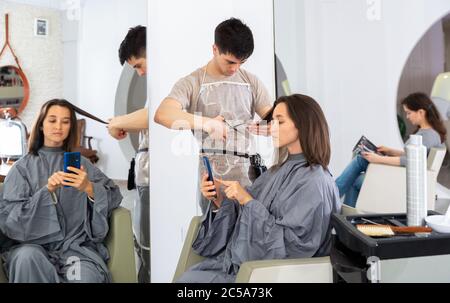 Positive male hairdresser making hair styling for young woman, woman using mobile phone Stock Photo
