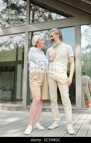 Happy beautiful senior couple in summer outfits looking at each other while standing against country house Stock Photo